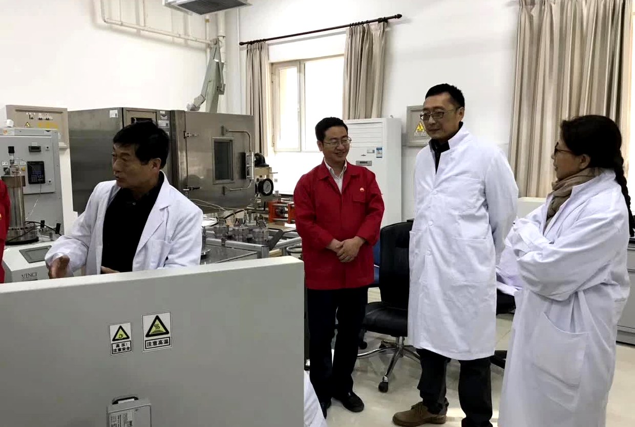 一位实验室技术人员站在仪器前讲解，四人在旁围听；前景白色挡板上贴有高压注意安全与注意高温两枚黄色警示签